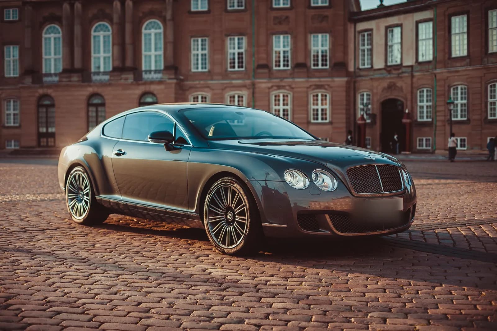 A dark Bentley Continental GT parked on a cobblestone street in front of a brick building.