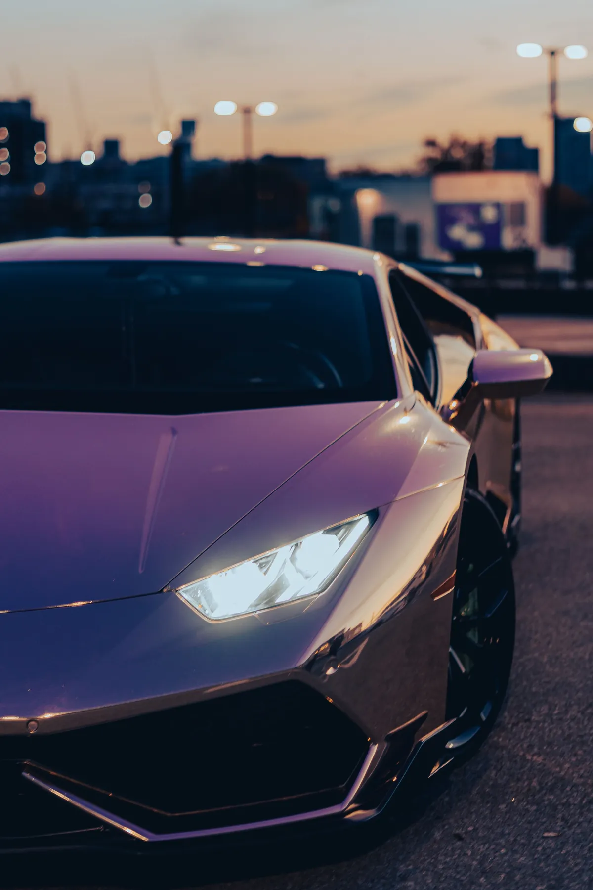 The front of a supercar at dusk, headlights lit, with city lights and a crane on the skyline behind it.