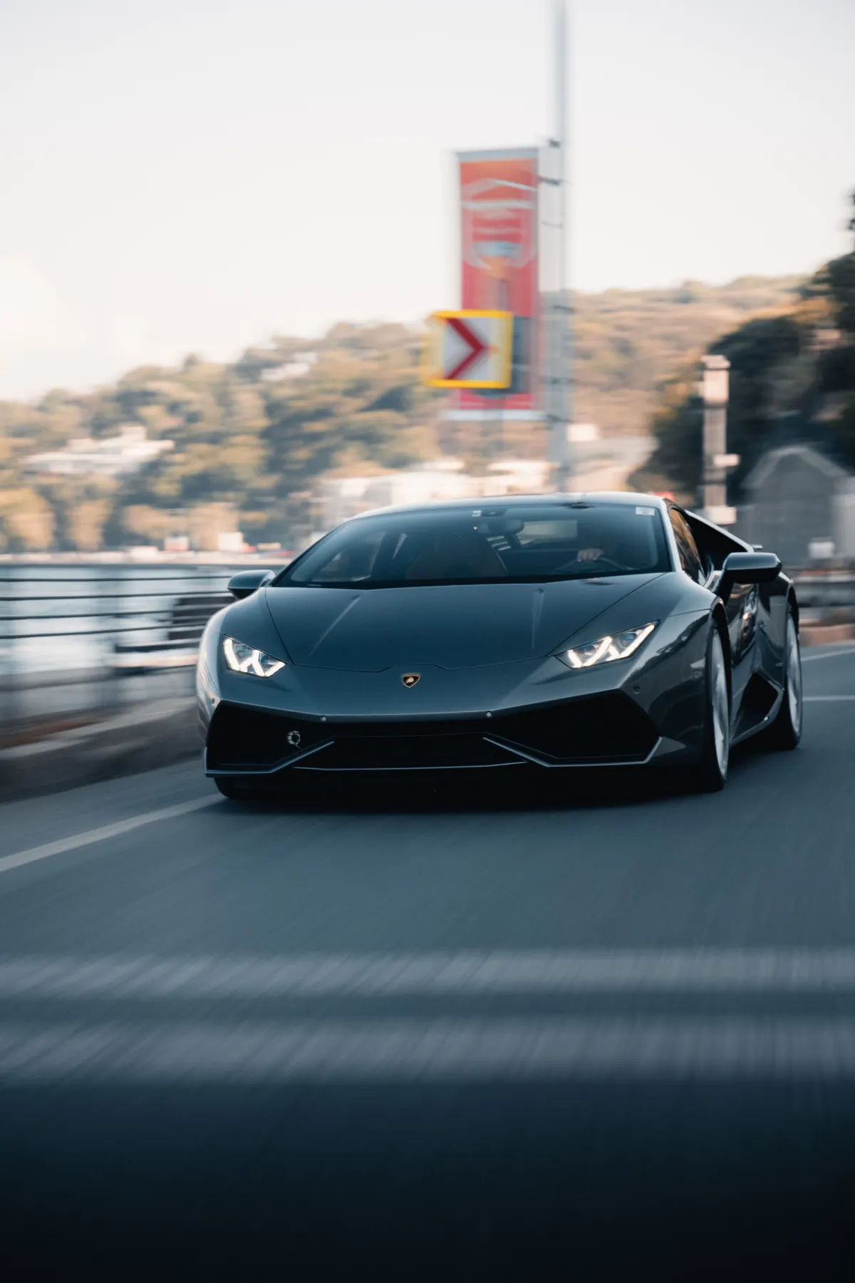 A dark supercar head-on at speed, the road and the guardrail blurred past it under a pale sky.